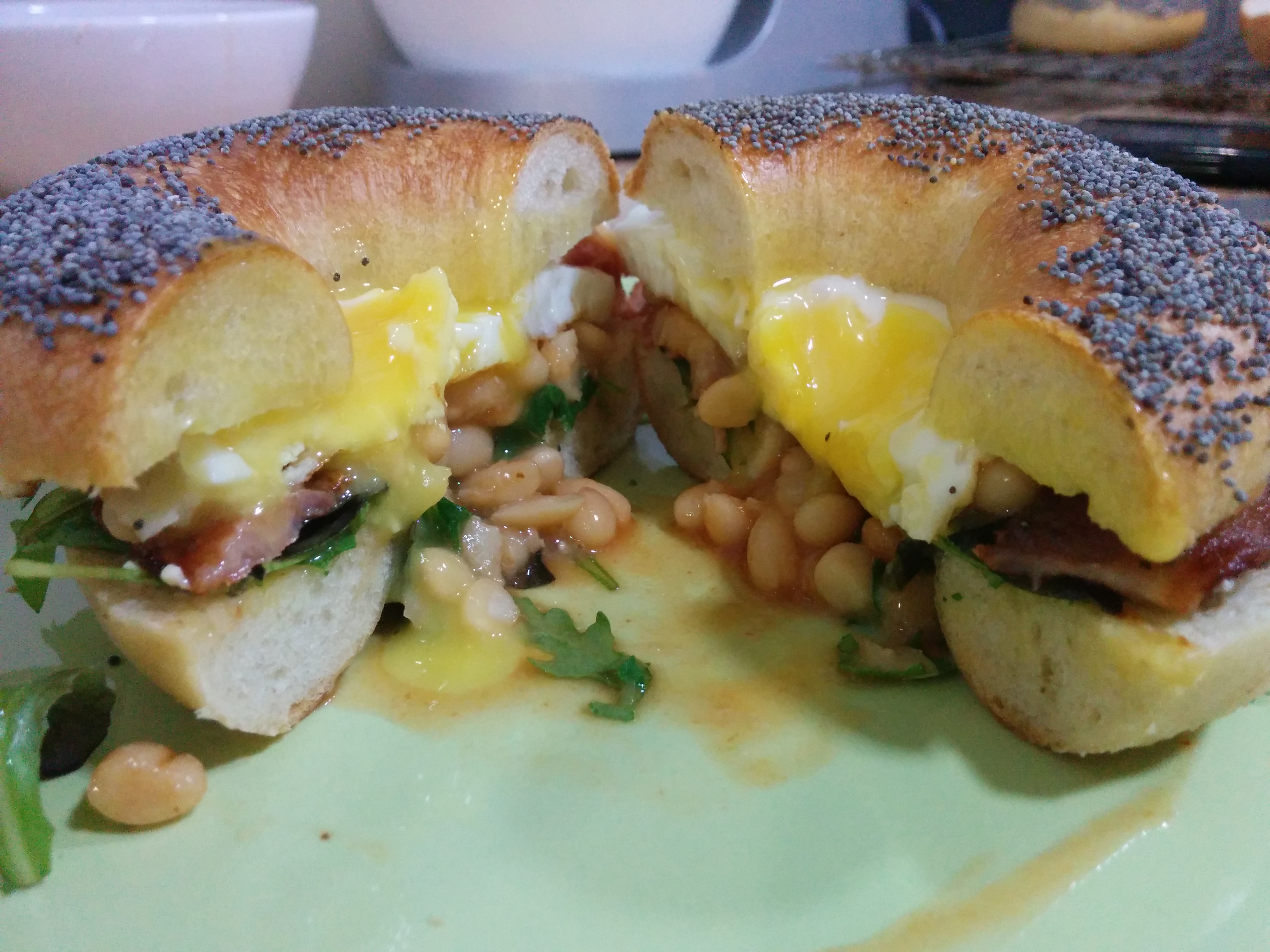 A bagel with rocket, fried backon, beans in tomato sauce and a fried egg